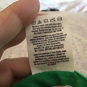 May include: A white clothing tag with care instructions in English, Spanish, and French. The tag includes washing symbols and text about washing with cold water, using non-chlorine bleach, and tumble drying. The tag is attached to a green fabric.