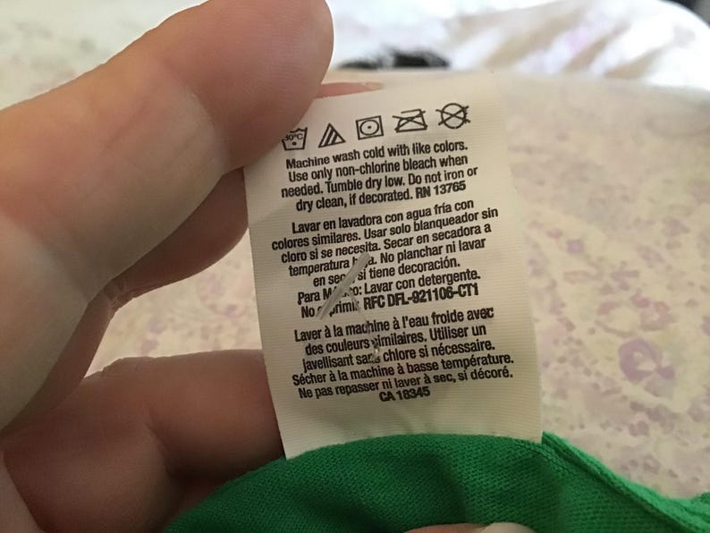 May include: A white clothing tag with care instructions in English, Spanish, and French. The tag includes washing symbols and text about washing with cold water, using non-chlorine bleach, and tumble drying. The tag is attached to a green fabric.