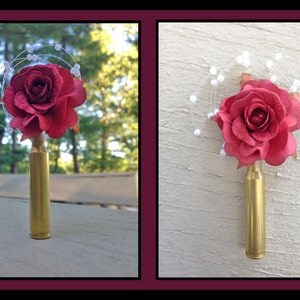 May include: A red rose with white pearl accents is attached to a gold bullet casing. The rose is on a wooden surface.