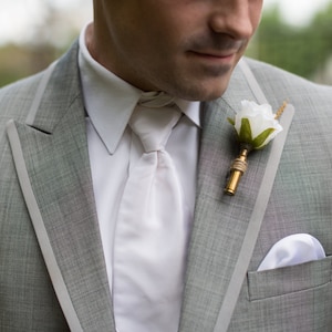 May include: A gray suit jacket with a white shirt and a white flower boutonniere. The boutonniere is attached to a gold pin.