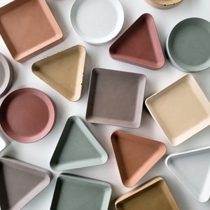 May include: Assortment of decorative trays in various shapes and colors, including round, square, and triangular. The trays are made of a textured material and have a rustic, minimalist aesthetic.