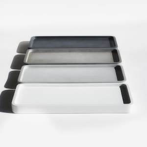 May include: Four rectangular trays made of concrete with black rubber edges. The trays are stacked on top of each other, with the darkest gray tray on top and the lightest white tray on the bottom.
