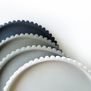 May include: Three round ceramic trays with scalloped edges. The trays are stacked, with the largest tray on the bottom and the smallest tray on top. The trays are in shades of gray, from dark gray to light gray.