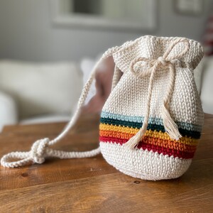 May include: A white crocheted drawstring bag with a rainbow stripe design and a long strap. The bag is made with a chunky yarn and has a simple, minimalist style.