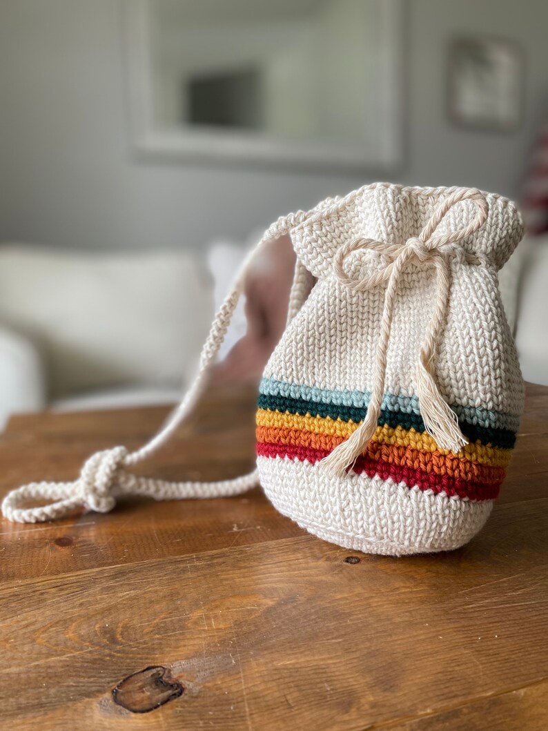May include: A white crocheted drawstring bag with a rainbow stripe design and a long strap. The bag is made with a chunky yarn and has a simple, minimalist style.
