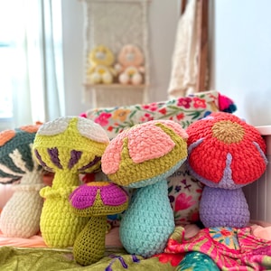 May include: Five crocheted mushroom-shaped plushies in various colors, including pink, green, yellow, blue, and red. The plushies are arranged on a colorful floral patterned fabric.