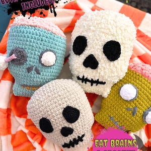 May include: Four crocheted skull-shaped pillows in different colors, including blue, white, gray, and yellow. The pillows are arranged on a checkered orange and white blanket. The text "Eat Brains & Crochet Pillow Crochet Patterns" is visible in the bottom right corner of the image.