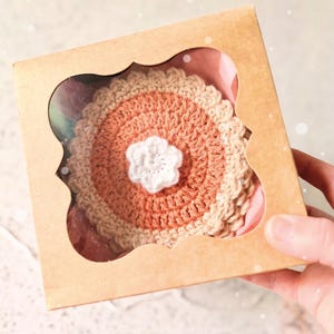 Puede incluir: Un juego de posavasos redondos de ganchillo en tonos terracota, beige y crema, con un pequeño detalle de flor blanca en el centro. Los posavasos están empaquetados dentro de una caja de cartón marrón claro con una ventana decorativa.