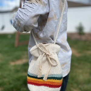 May include: A white crocheted drawstring bag with a rainbow stripe design. The bag is worn as a crossbody bag.
