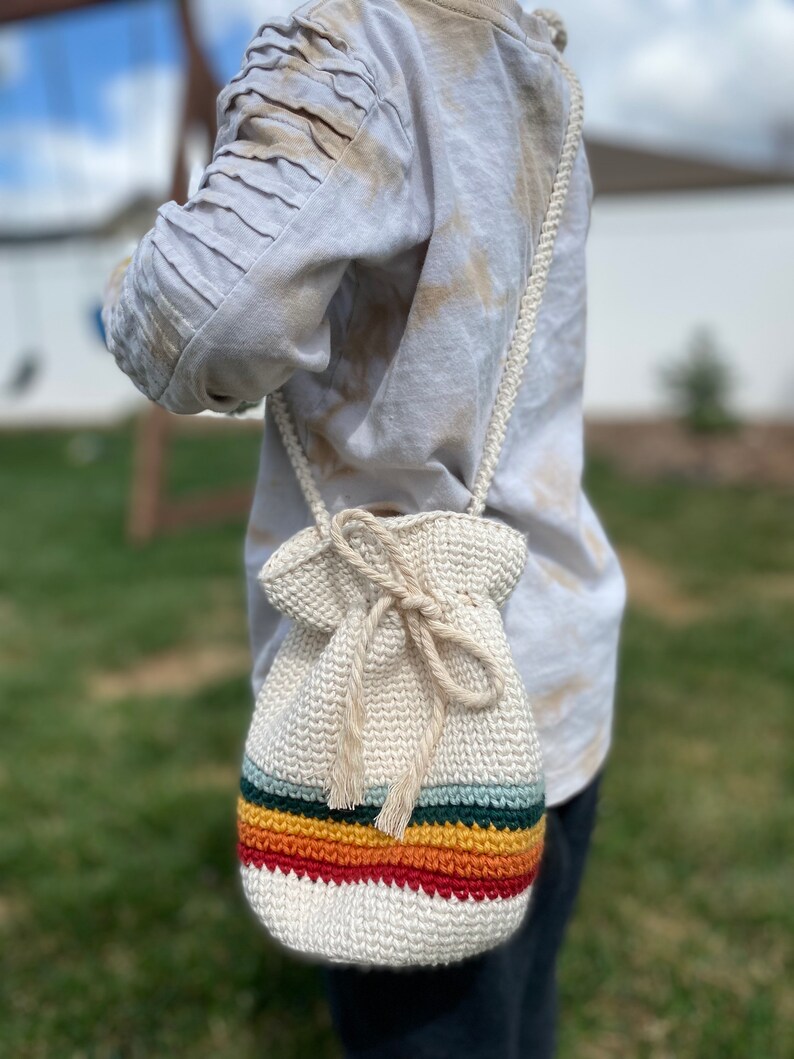 May include: A white crocheted drawstring bag with a rainbow stripe design. The bag is worn as a crossbody bag.