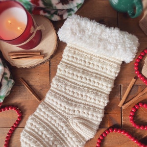 May include: A white crocheted Christmas stocking with a fluffy white cuff. The stocking is decorated with a textured pattern and is lying on a wooden surface with red beads, cinnamon sticks, and a lit candle.