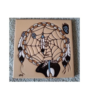 May include: A square ceramic tile featuring a raised relief design of a dreamcatcher with a central web, hanging feathers, and a bear silhouette at the bottom. The color palette includes tan, brown, black, white, and light blue accents with a signature.