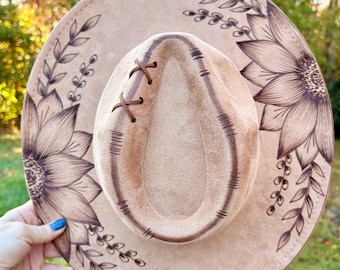 Pode incluir: Chapéu bege com aba larga, com design floral e detalhes costurados. A aba é decorada com grandes girassóis e vinhas frondosas. A copa tem um design costurado com detalhes de couro cruzados.
