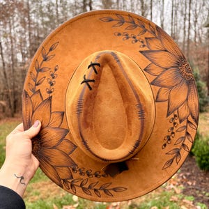 Pu&ograve; includere: Un cappello a tesa larga color marrone chiaro con illustrazioni a linee nere raffiguranti girasoli e rami frondosi. La corona presenta dettagli di cuciture nere e un piccolo accento a punto croce sulla parte anteriore.