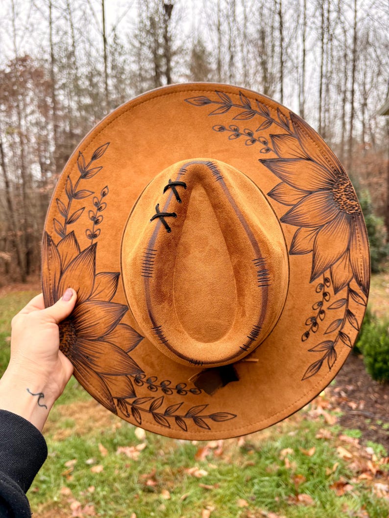 Pu&ograve; includere: Un cappello a tesa larga color marrone chiaro con illustrazioni a linee nere raffiguranti girasoli e rami frondosi. La corona presenta dettagli di cuciture nere e un piccolo accento a punto croce sulla parte anteriore.