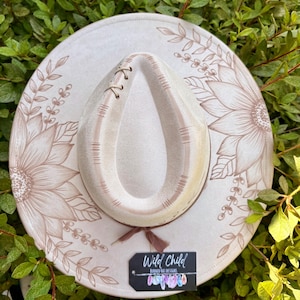 Pu&ograve; includere: Un cappello a tesa larga beige chiaro con illustrazioni floreali a linee marroni sulla tesa, una corona a forma di goccia con cuciture decorative e un nastro marrone. Un'etichetta rettangolare nera riporta la scritta Wild Child Burned Hat Designs.