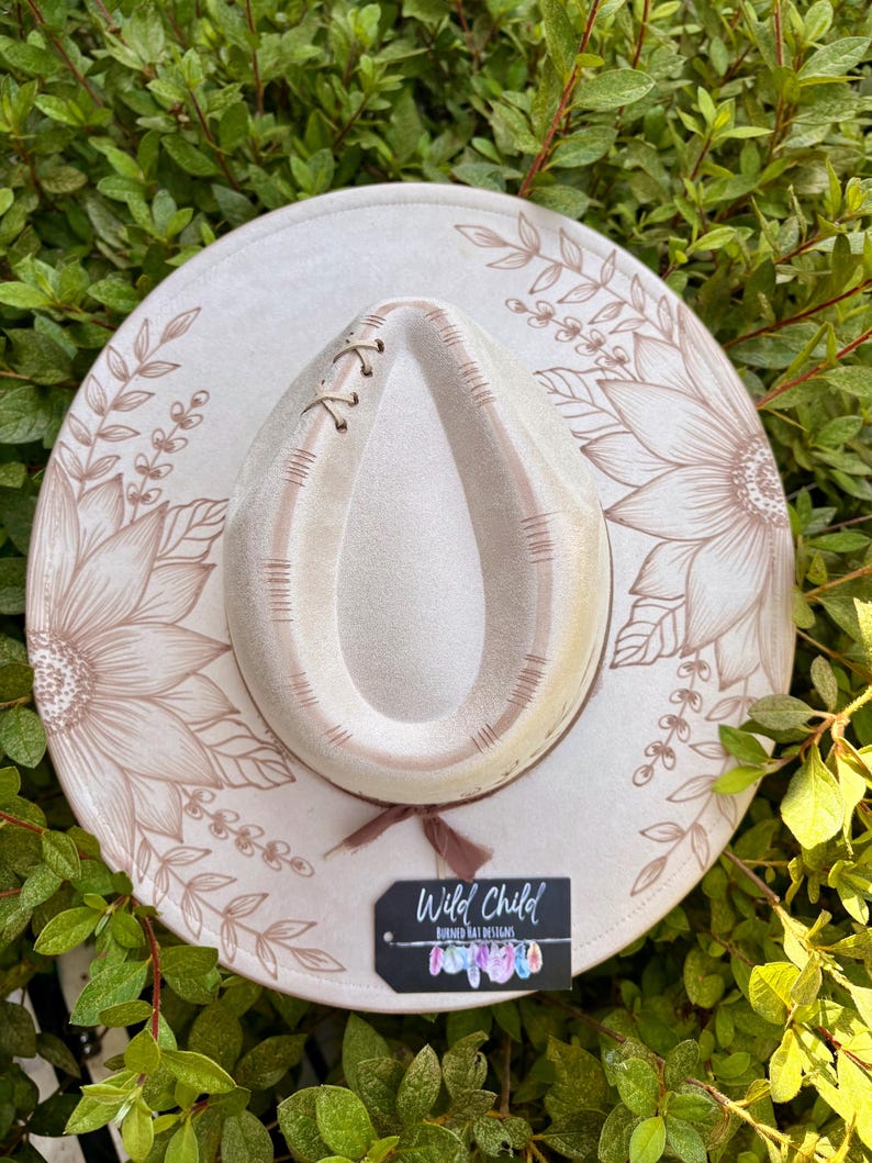 Pu&ograve; includere: Un cappello a tesa larga beige chiaro con illustrazioni floreali a linee marroni sulla tesa, una corona a forma di goccia con cuciture decorative e un nastro marrone. Un'etichetta rettangolare nera riporta la scritta Wild Child Burned Hat Designs.