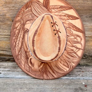 May include: A tan leather hat with a wide brim, featuring an embossed Native American design with feathers. The hat has a teardrop-shaped crown with brown lacing. The tag reads "Wild Child, Burned Hat Designs."