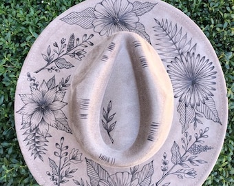 May include: A light brown felt fedora hat with a black floral design. The hat has a wide brim and a tall crown. The design features various flowers, leaves, and stems. The hat has a tag that says "Wild Child".