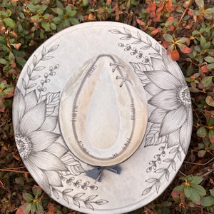 Pu&ograve; includere: Un cappello a tesa larga grigio chiaro con un disegno floreale a inchiostro nero disegnato a mano, raffigurante grandi girasoli e tralci di foglie. La corona ha una forma a goccia con cuciture decorative e un nastro grigio.