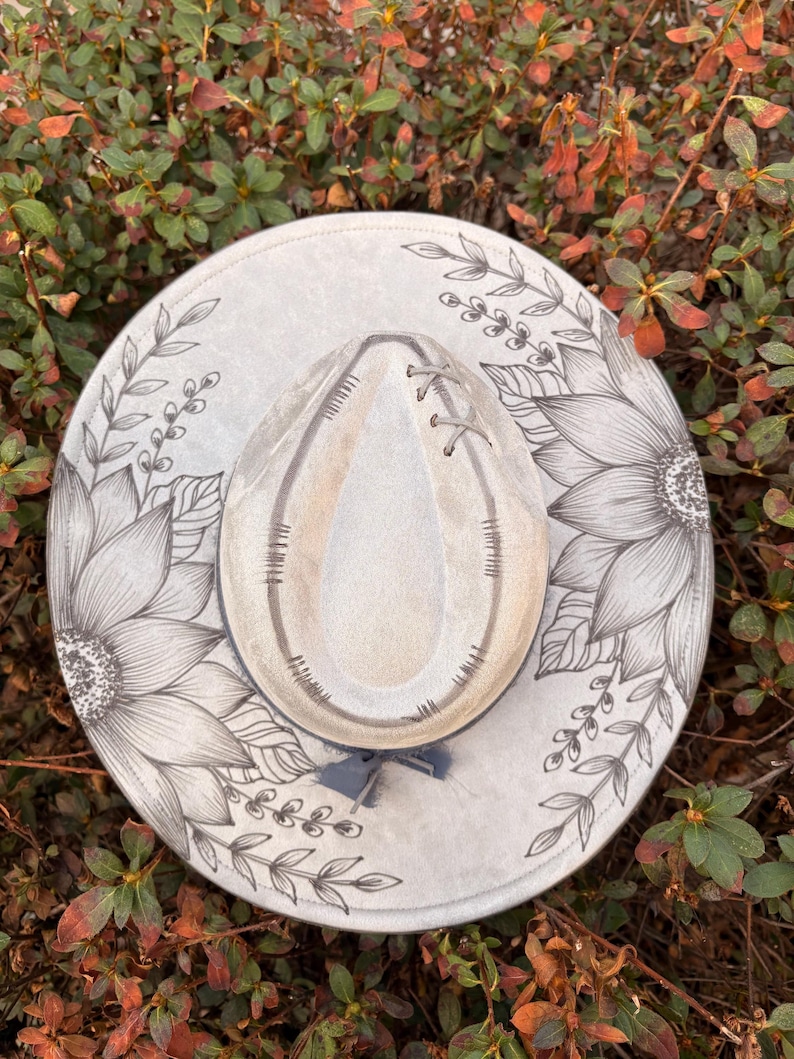 Pu&ograve; includere: Un cappello a tesa larga grigio chiaro con un disegno floreale a inchiostro nero disegnato a mano, raffigurante grandi girasoli e tralci di foglie. La corona ha una forma a goccia con cuciture decorative e un nastro grigio.