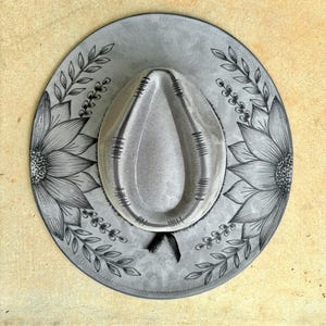 Pu&ograve; includere: Un cappello in feltro grigio con una corona a forma di goccia e decorativi accenti cuciti in nero. La tesa larga &egrave; ornata con illustrazioni a inchiostro nero disegnate a mano di grandi girasoli e motivi di viti frondose sui lati.
