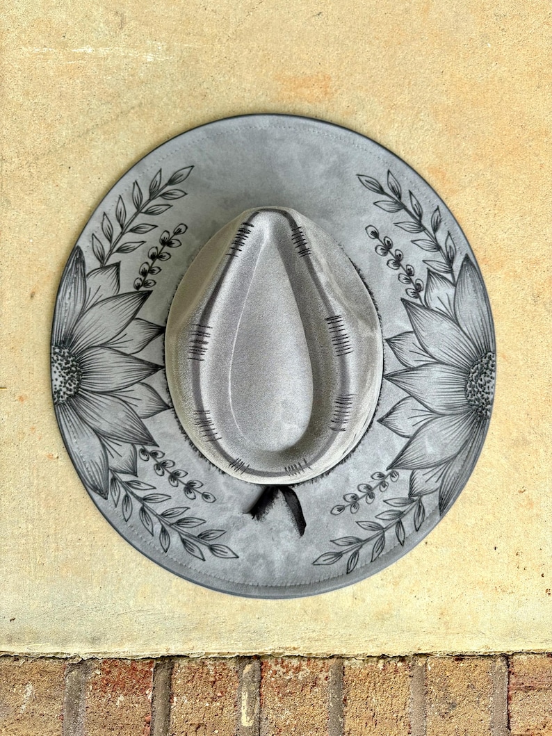 Pu&ograve; includere: Un cappello in feltro grigio con una corona a forma di goccia e decorativi accenti cuciti in nero. La tesa larga &egrave; ornata con illustrazioni a inchiostro nero disegnate a mano di grandi girasoli e motivi di viti frondose sui lati.