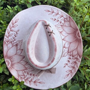 Pu&ograve; includere: Un cappello rosa chiaro con tesa larga decorata con illustrazioni a linee rosa scuro di girasoli e tralci di foglie. La corona presenta un'indentazione a goccia con dettagli a punto croce marrone scuro e un piccolo fiocco.