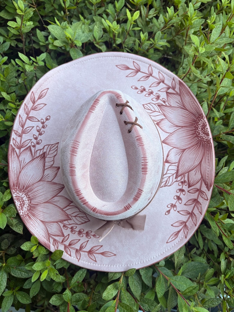 Pu&ograve; includere: Un cappello rosa chiaro con tesa larga decorata con illustrazioni a linee rosa scuro di girasoli e tralci di foglie. La corona presenta un'indentazione a goccia con dettagli a punto croce marrone scuro e un piccolo fiocco.