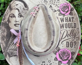 Può includere: Un cappello beige a tesa larga con il ritratto di una donna, rose rosa e il testo "WHAT WOULD DOLLY DO?". Il cappello è decorato con note musicali, cuori, un nastro rosa e una piuma marrone, con cuciture rosa sulla corona.