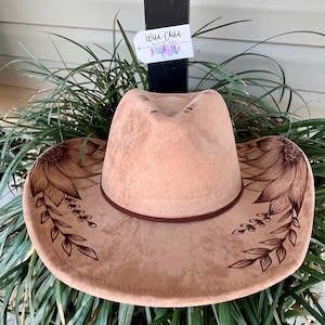 Pu&ograve; includere: Un cappello da cowboy in feltro color marrone chiaro con disegni a linee marrone scuro di girasoli e viti frondose sulla tesa. Una fascia marrone scuro circonda la base della corona. Un'etichetta rettangolare bianca con il testo Wild Child.