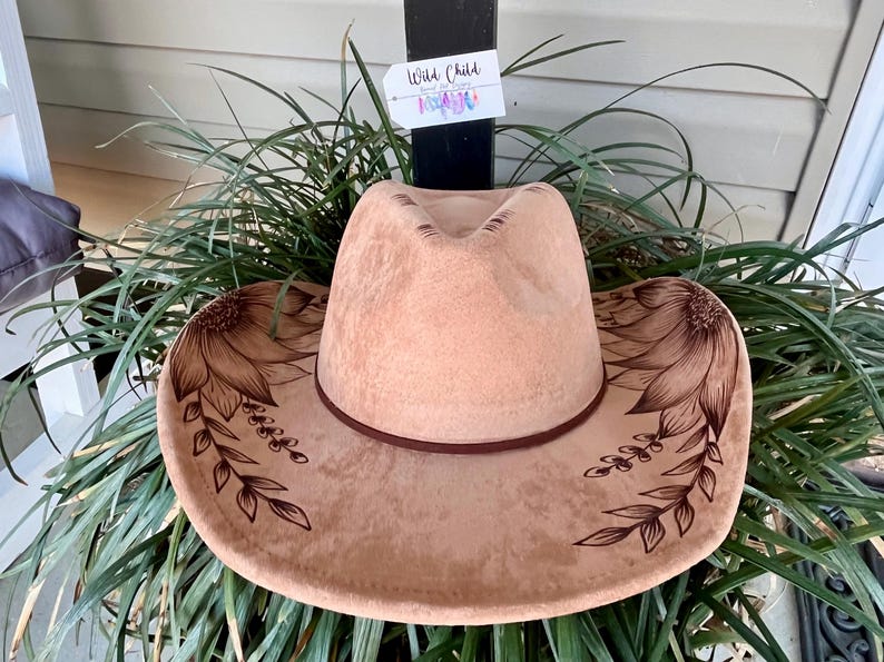 Pu&ograve; includere: Un cappello da cowboy in feltro color marrone chiaro con disegni a linee marrone scuro di girasoli e viti frondose sulla tesa. Una fascia marrone scuro circonda la base della corona. Un'etichetta rettangolare bianca con il testo Wild Child.