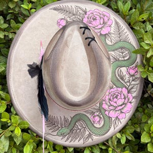 May include: A wide-brimmed beige hat featuring a detailed illustration of a green snake winding through pink peony flowers and fern leaves. The crown is accented with black stitching and a decorative arrangement of black and pink feathers on the side.