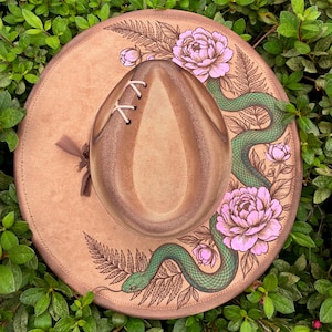 May include: A tan wide-brimmed hat featuring a hand-drawn illustration of a green snake winding through pink peony flowers and fern leaves. The crown includes a decorative white lace-up detail on one side and a small brown ribbon tie.