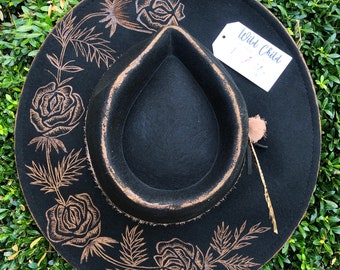 May include: Black felt fedora hat with a gold-colored design of roses and leaves. The hat has a wide brim and a feather accent. The hat has a tag that says "Wild Child".