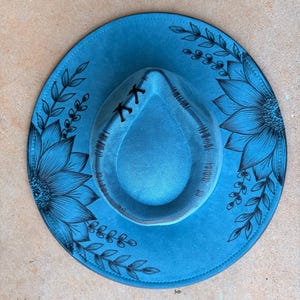 Pu&ograve; includere: Un cappello a tesa larga di colore blu brillante con illustrazioni a linee nere di grandi girasoli e viticci frondosi sulla tesa. La corona del cappello presenta dettagli cuciti neri e un piccolo accento con lacci neri sul davanti.