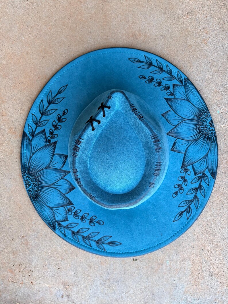 Pu&ograve; includere: Un cappello a tesa larga di colore blu brillante con illustrazioni a linee nere di grandi girasoli e viticci frondosi sulla tesa. La corona del cappello presenta dettagli cuciti neri e un piccolo accento con lacci neri sul davanti.