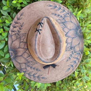 Pu&ograve; includere: Un cappello a tesa larga color marrone chiaro con illustrazioni a inchiostro marrone scuro di grandi girasoli e viti frondose sulla tesa. La corona presenta un'indentazione centrale con cuciture nere e un nastro nero alla base.