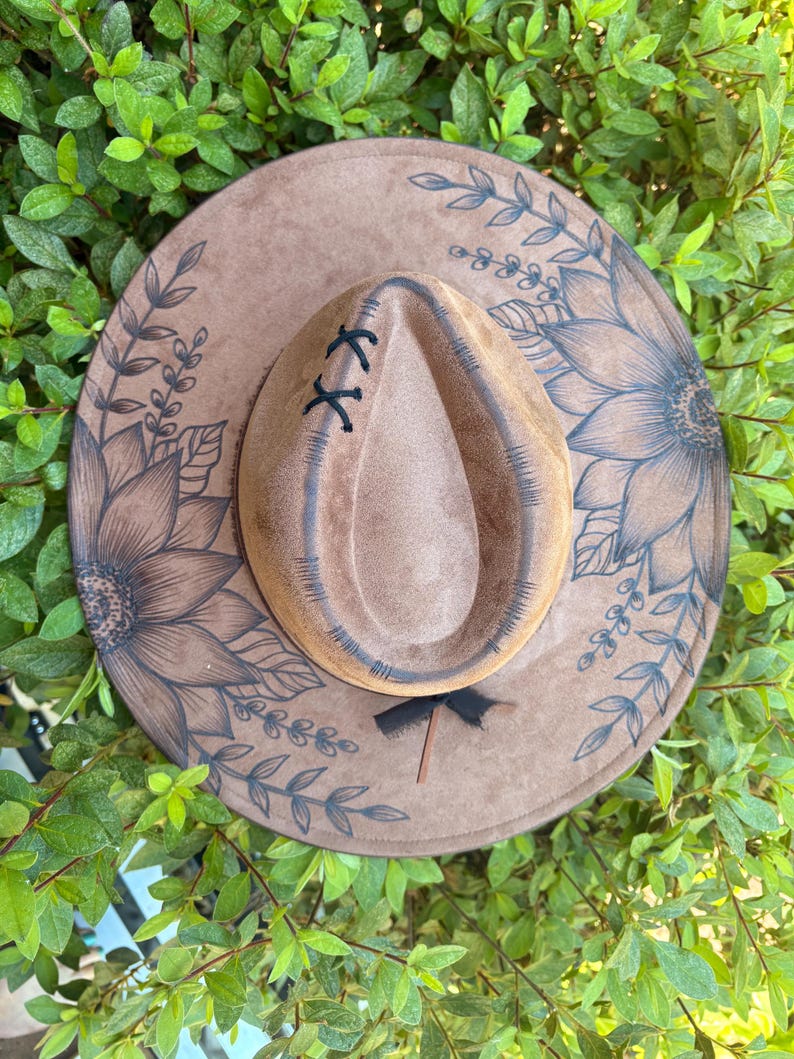 Pu&ograve; includere: Un cappello a tesa larga color marrone chiaro con illustrazioni a inchiostro marrone scuro di grandi girasoli e viti frondose sulla tesa. La corona presenta un'indentazione centrale con cuciture nere e un nastro nero alla base.
