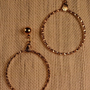May include: A pair of gold-colored hoop earrings. Each earring features a small, round stud at the top, connected to a large, textured hoop. The hoops have a twisted rope design. The earrings are displayed against a neutral, textured background.