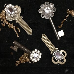 Könnte beinhalten: Vier dekorative Schlüssel mit Juwelenakzenten. Jeder Schlüssel hat ein anderes Design und verfügt über einen klaren Kristall-Schmuck. Die Schlüssel sind auf einer schwarzen Oberfläche angeordnet.