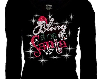 May include: A black long-sleeve shirt featuring a rhinestone graphic on the chest. The design includes the text "Bling it on Santa" in white, green, and red crystals, accented with a small Santa hat, snowflake shapes, and radiating starbursts.