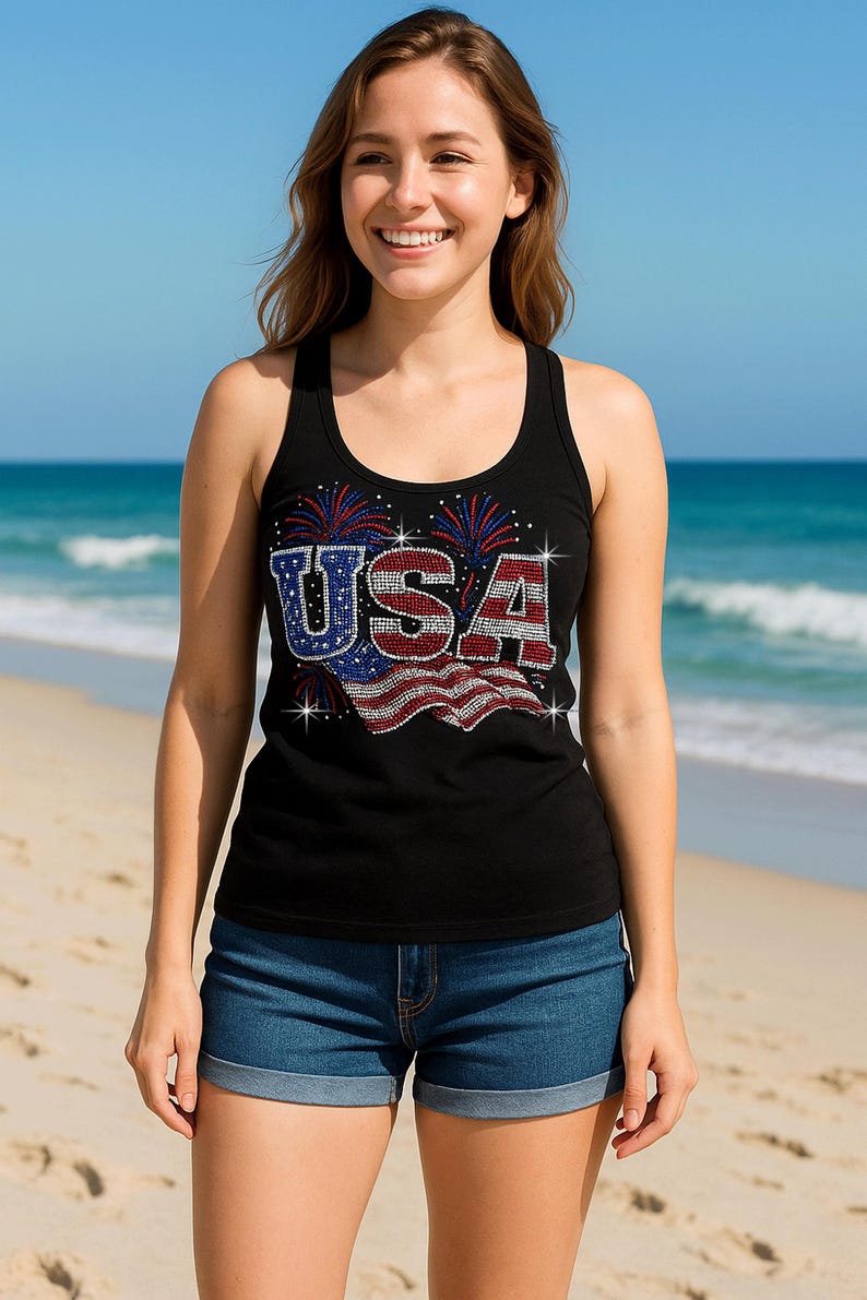 May include: Black tank top with the word "USA" in a patriotic design, featuring red, white, and blue accents. The tank top is paired with denim shorts. The background shows a beach and ocean.