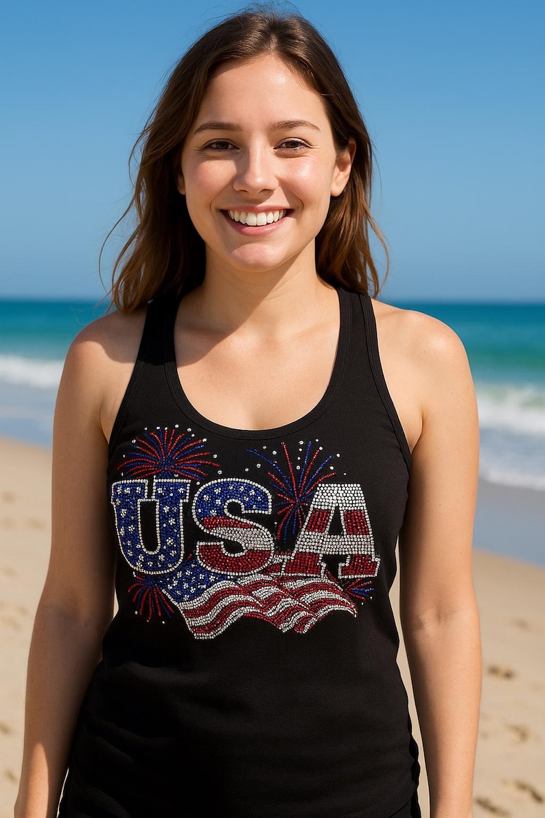 May include: Black tank top with a patriotic "USA" design in red, white, and blue rhinestones, featuring a waving flag and fireworks. The top is sleeveless and has a scoop neck.