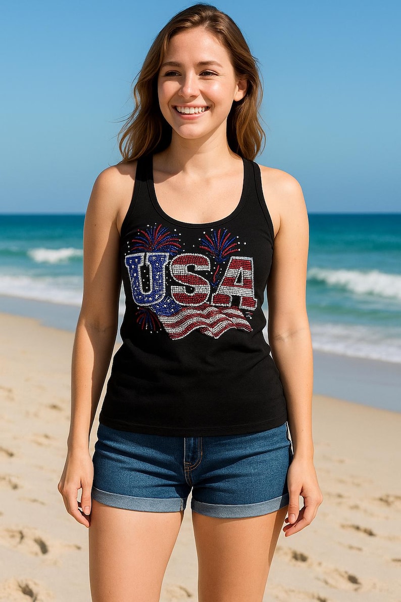 May include: Black tank top with the word "USA" in red, white, and blue rhinestones. The tank top has a scoop neck and is paired with denim shorts. The background is a beach with blue water and sky.