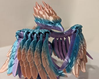 May include: A 3D printed articulated bird figurine featuring a gradient color scheme of metallic peach, bright blue, and purple. The creature has a sharp beak, detailed layered wing feathers, and a spiky crest on its head, all constructed with visible horizontal print layers.