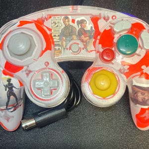 May include: A clear GameCube controller with red and white splatter design. The controller features images of characters from Resident Evil and the text "RESIDENT EVIL". It has gray, green, yellow, and red buttons, and a black cable.