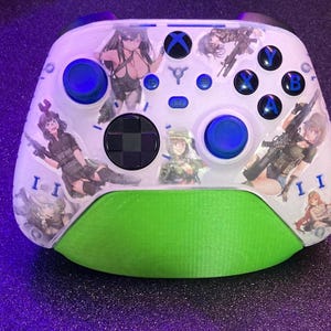 May include: Custom white Xbox controller with a green base and blue buttons. The controller is decorated with anime-style illustrations of women holding weapons. The buttons are labeled with the letters A, B, X, and Y.