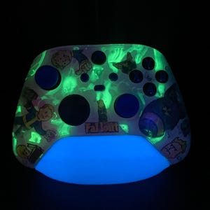 May include: A custom video game controller with a Fallout theme. The controller has a glowing blue base and green accents. It features character illustrations and the word "Fallout". The buttons are blue.