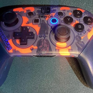 May include: A Nintendo Switch Pro Controller with a custom design. The controller features a blue and gray color scheme with orange accents. The design includes images of characters and the text "Umbrella".